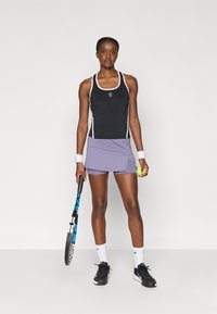 Fekete sportos tanktop fehér díszítéssel, lila szoknyával párosítva. A modell teniszütőt és egy labdát tart, fekete tornacipőt visel.