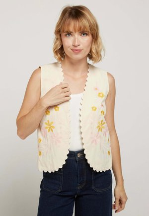 Femme aux cheveux blonds portant un gilet crème à bords festonnés avec une broderie florale jaune et rose, sur un haut blanc et un jean foncé.