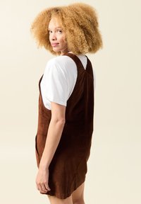 Braunes Corduroy-Latzkleid mit figurbetoneter Form und seitlichen Taschen. Kombiniert mit einem weißen T-Shirt, das den strukturierten Stoff und die verstellbaren Träger zeigt.