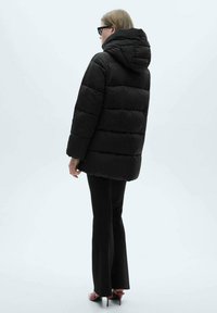 Jachetă puf din material negru, cu glugă, având un design matlasat și o croială lejeră. Purtați-o cu pantaloni negri și pantofi cu toc înalt roșii.