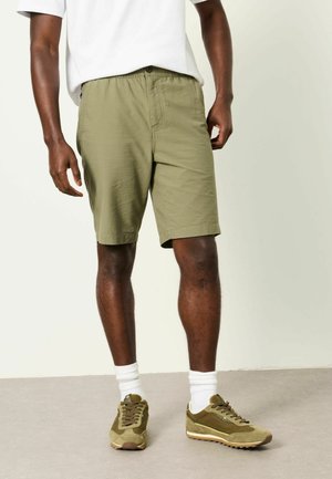 Person trägt olivgrüne, strukturierte Shorts, weiße Socken, olivgrüne Sneakers und ein weißes T-Shirt vor neutralem Hintergrund.