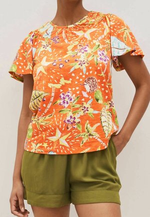 Femme portant un haut orange à manches courtes avec un imprimé tropical multicolore et un short vert olive, debout avec une main dans la poche.