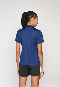 Mornarsko polo majica kratkih rukava s ovratnikom i žutim logotipom na rukavu. Tkanina ima glatku teksturu, nosi se s crnim sportskim kratkim hlačama.