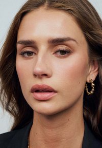 Guldörhängen med sfäriska accenter, slät hud, naturlig sminkning och långt, vågigt brunt hår. Neutral bakgrund.