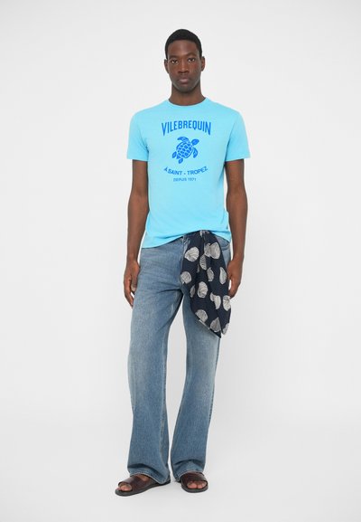 Moški v svetloplavem majici s potiskom, ohlapnih modrih kavbojkah, s črno-belim vzorčastim šalom in temno rjavimi sandali, stoji ob belem ozadju.