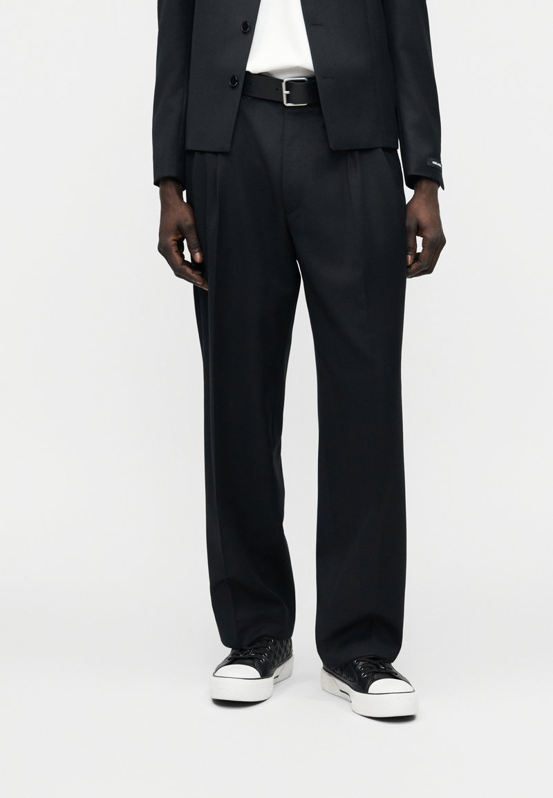 Άνδρας που φοράει μαύρο πλισέ παντελόνι, μαύρη ζώνη, μαύρο σακάκι, λευκό πουκάμισο και μαύρα και λευκά αθλητικά παπούτσια, στέκεται μπροστά σε απλό φόντο.