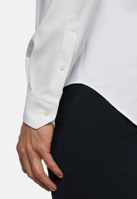 Chemise blanche à manches longues, avec une texture lisse, des poignets boutonnés et une coupe ajustée. Tissu sombre visible en dessous.