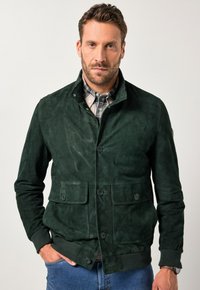 Giacca in suede verde con colletto alto, due tasche frontali e bottoni neri. Indossata sopra una camicia a quadri e abbinata a dei jeans.