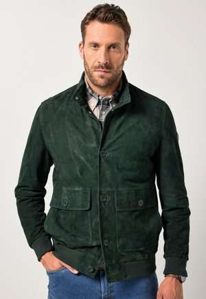 Chaqueta de ante verde con un alto cuello, dos bolsillos delanteros y botones negros. Llevada sobre una camisa a cuadros y combinada con denim.