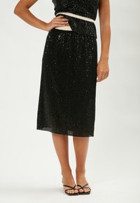 Falda midi negra de lentejuelas con textura plisada, cinturilla elástica y dobladillo en capas. Combinada con sandalias negras de tiras.