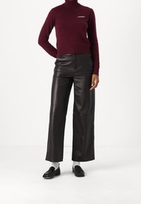 Osoba ubrana w bordowy sweter z golfem z napisem "2NDDAY", czarne szerokie skórzane spodnie, białe skarpetki i czarne loafersy na białym tle.