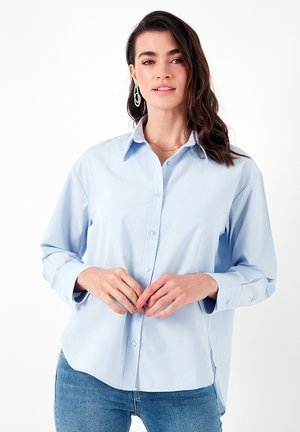 Light blue long-sleeve button-up shirt with a collar, featuring a relaxed fit and smooth cotton fabric. Worn with mid-rise jeans.