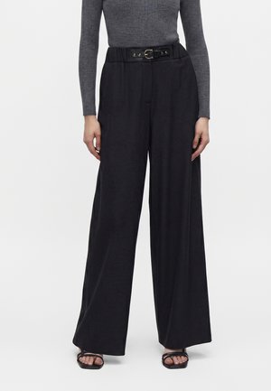 Femme portant un pantalon large noir taille haute avec une ceinture noire, un haut gris ajusté à manches longues et des sandales noires ouvertes à l'avant.