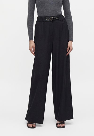 Femme portant un pantalon large noir taille haute avec une ceinture noire, un haut gris ajusté à manches longues et des sandales noires ouvertes à l'avant.