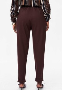 Pantalons marron extensibles avec une coupe ajustée, dotés de poignet à la cheville, portés avec des chaussures sombres à lanières. Texture de tissu douce.