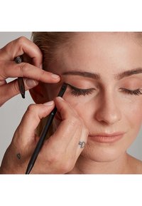 Applicazione del trucco in corso, con un primo piano del viso di un modello con trucco naturale. Mani che tengono una matita eyeliner nera vicino all'occhio.