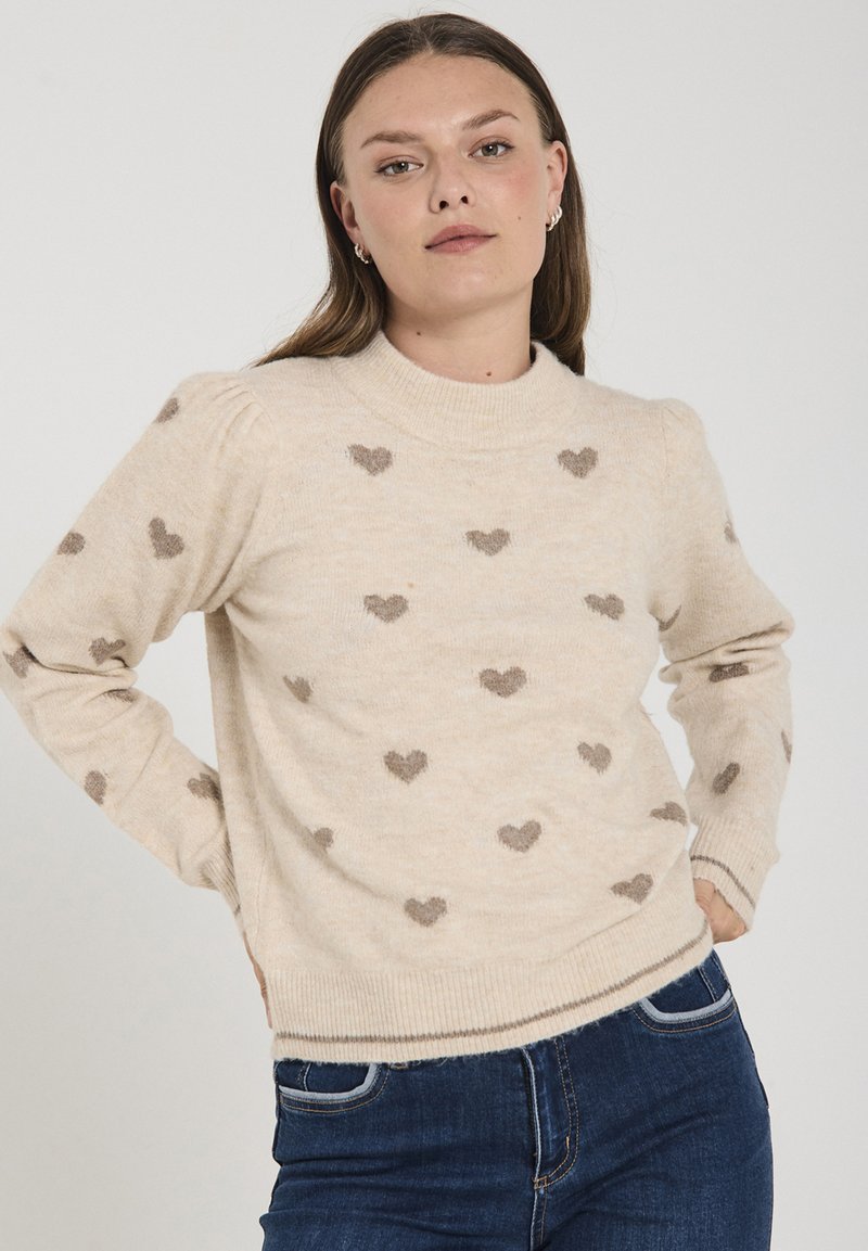 Beige stickad tröja med små bruna hjärtan, ribbad rundad halsringning och åtsittande ärmar. Mjuk textur, avslappnad passform och lätt puffade axlar.
