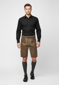 Lederhosen de cuero marrón con costuras decorativas, combinados con una camiseta de manga larga negra y calcetas gris de longitud hasta la rodilla, terminados con zapatos negros.