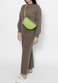 Borsa a tracolla verde in pelle con un design curvato, indossata sopra un maglione corto marrone e pantaloni a gamba larga coordinati. Infradito chiari.