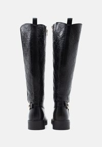 Vysoké černé kožené boty s embosovanými kruhovými vzory, bočními zipy a kotníkovými pásky se zlatými přezkami, zobrazené zezadu.