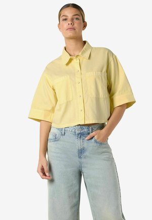 Žena nosi krátku žltú košeľu na gombíky s dvoma náprsnými vreckami a svetlomodré rifle s vysokým pásom, pózuje s jednou rukou v vrecku.