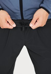 Zwarte broek met een elastische tailleband, gladde textuur, gedeeltelijk bedekt door handen die de tailleband vasthouden. Grijze top zichtbaar erboven.
