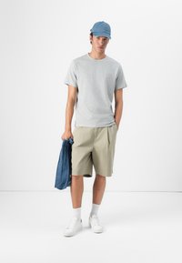 Grå t-skjorte med rund hals, beige shorts, hvite sneakers og blå denim caps. Holder en blå denimjakke, står mot en hvit bakgrunn.