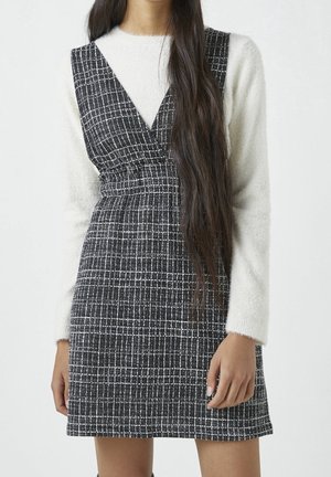 Femme portant une robe sans manches à carreaux noirs et blancs par-dessus un haut blanc en tissu duveteux à manches longues, avec de longs cheveux foncés tombant sur une épaule.