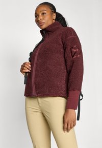 Bordeaux fleece jakke med høy krage, teksturert overflate, svart glidelås og justerbare snorer i kanten, med brodert logo på ermet.