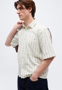 Νέος άνδρας που φοράει μια κοντομάνικη μπεζ πουκαμίσα με κάθετες ρίγες, κρατώντας ένα καφέ σακάκι στον ώμο του, με το χέρι στην τσέπη του τζιν.