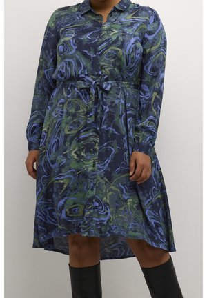 Langärmliges Hemdkleid mit blau-grünem abstraktem Muster, Taillengürtel und High-Low-Saum, kombiniert mit schwarzen kniehohen Stiefeln.