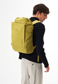 Gelbes Rucksack mit verstellbarem Oberteil, rechteckiger Form, Fronttasche und zwei runden Akzenten. Strukturierter Stoff, wird über einer Schulter getragen.