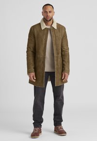 Brauner Wildledermantel mit einem Schafslederkragen, langen Ärmeln und einem vorderen Verschluss, kombiniert mit grauen Hosen und braunen Stiefeln.