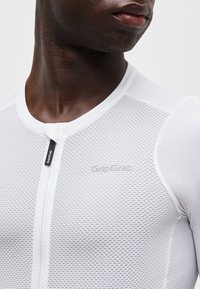 Κοντινό πλάνο ενός ατόμου που φοράει ένα λευκό ποδηλατικό μπλουζάκι με υφή, με φερμουάρ μπροστά και το λογότυπο "GripGrab" στο στήθος.