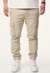 Beige Cargo-Hosen mit elastischen Bündchen, ausgestattet mit seitlichen Taschen und einem Kordelzugbund. Getragen mit weißen Sneakers, entworfen für legeres Tragen.
