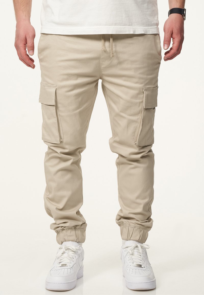 Beige Cargo-Hosen mit elastischen Bündchen, ausgestattet mit seitlichen Taschen und einem Kordelzugbund. Getragen mit weißen Sneakers, entworfen für legeres Tragen.