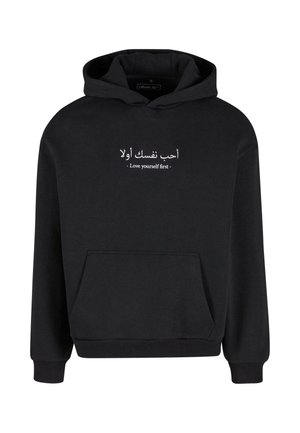 Zwarte hoodie gemaakt van zachte stof, met een voorvak. Tekst in witte Arabische en Engelse letters leest "Hou eerst van jezelf." Verstelbare capuchon.