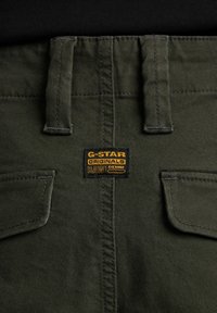 Olivinės spalvos kelnės iš tekstūruotos medžiagos, su dviem nugaros kišenėmis ir G-Star RAW džinsiniu ženklu, siuvinėtu ant liemens.