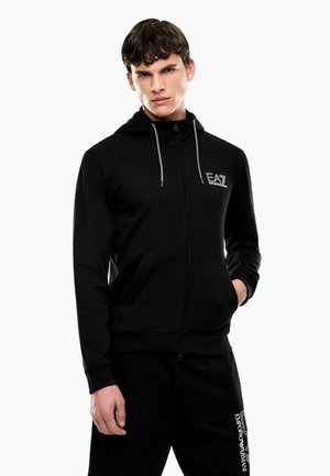 Młody mężczyzna ubrany w czarną bluzę z kapturem i zamkiem EA7 Emporio Armani oraz czarne spodnie dresowe z logo marki, stojący z jedną ręką w kieszeni.