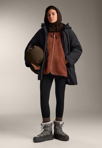 Doudoune noire, sweat zippé marron en polaire avec col montant, leggings noirs et bottes d'hiver grises avec doublure en fourrure. Porte un sac marron.