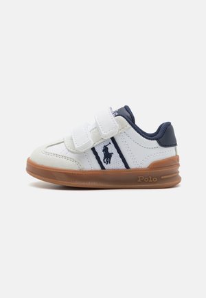 White sneaker with navy accents, featuring two Velcro straps, a textured suede toe, and a gum sole; branded with a logo on the side.