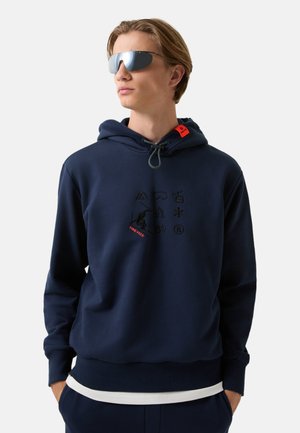 Junger Mann trägt reflektierende Sonnenbrille und einen dunkelblauen Hoodie mit schwarzen und roten grafischen Symbolen auf der Vorderseite, die Hände in den Taschen.