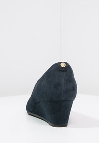 Scarpa con zeppa in suede blu scuro, con punta arrotondata e un piccolo accento metallico sul retro. Texture liscia con un design del tacco affusolato.