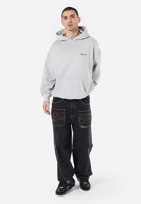 Sweatshirt cinza claro com um bolso canguru, combinado com calças de denim pretas de corte largo, com costuras laranja em contraste e bolsos decorativos.