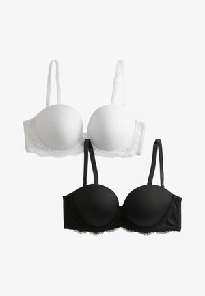 Vrouw draagt zwarte gewatteerde beha met kanten randjes en bijpassende zwarte bikini-onderbroek, staand tegen een effen lichte achtergrond.