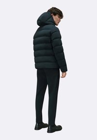 Giacca a vento nera con cappuccio, caratterizzata da un design trapuntato e forme cilindriche. Indossata con pantaloni scuri e scarpe nere.