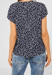 Kvinna som bär en svart kortärmad blus med vita prickar och ljusblå jeans, fotograferad bakifrån.