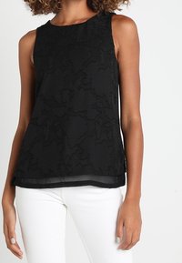 Svart ärmlös topp med texturerade blommönster, genomskinlig fåll och rund halsringning, kombinerad med vita byxor.