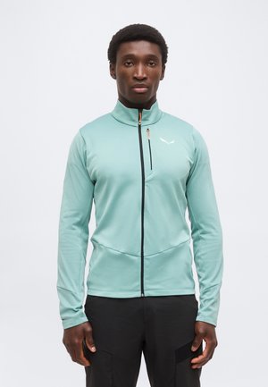 Mees, kes kannab helesinist lukuga jakki kõrge kaelusega ja musti pükse, seisab ühevärvilise valge tausta ees.