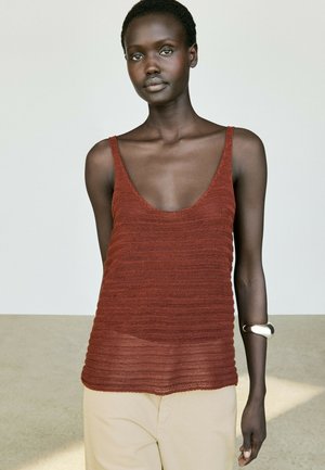 Femme aux cheveux courts portant un haut en tricot sans manches de couleur rouille et un pantalon beige, debout devant un fond clair uni.